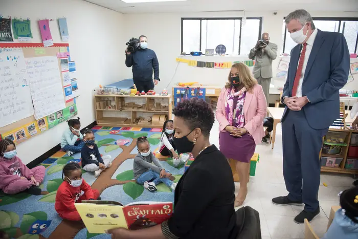 Mayor de Blasio and Chancellor Porter stand, while wearing masks, and watching kids in a 3K class sit on a mat while their teacher reads a book. Everyone is masked.